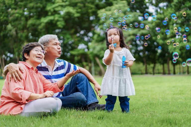 Asian granddaughter making bubbles and happy grandparents sitting in the grass