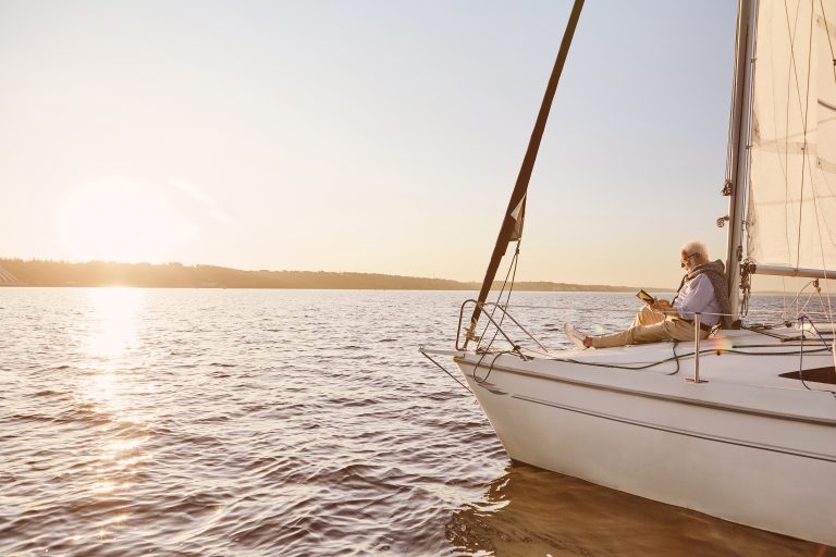 Sail boat floating in sea at sunset with senior man reading a book, sitting on the deck
